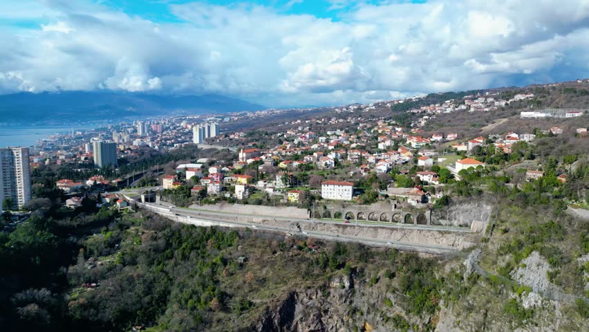 Aerial view of Rijeka, Croatia, showcasing its vibrant coastal urban landscape, port, and dramatic cloudy skies. A major road winds through the hills.
