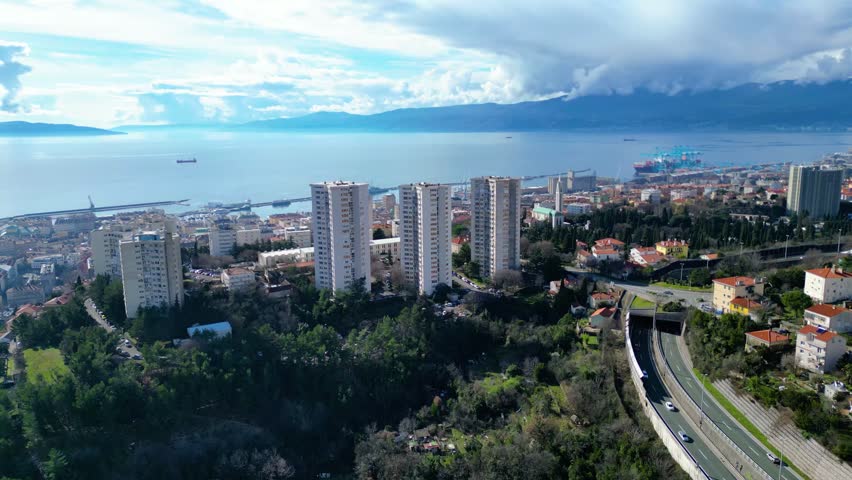 Aerial view of Rijeka, Croatia, showcasing its bustling port, diverse architecture, and the serene Adriatic Sea backed by distant mountains under a dynamic sky.