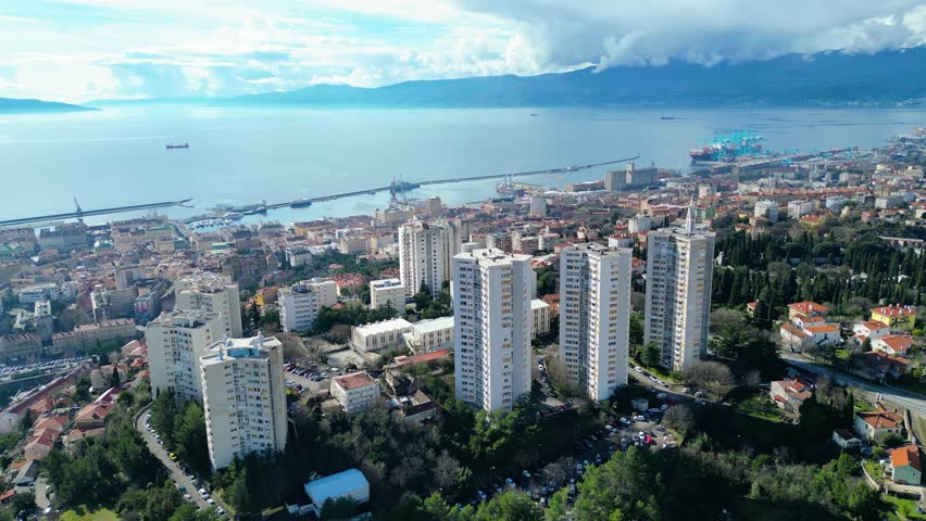 Aerial view of Rijeka, Croatia, showcasing its bustling port, diverse architecture, and the serene Adriatic Sea backed by distant mountains under a dynamic sky.