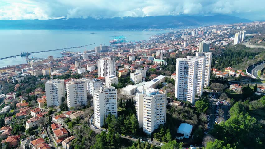 Aerial view of Rijeka, Croatia, showcasing its bustling port, diverse architecture, and the serene Adriatic Sea backed by distant mountains under a dynamic sky.