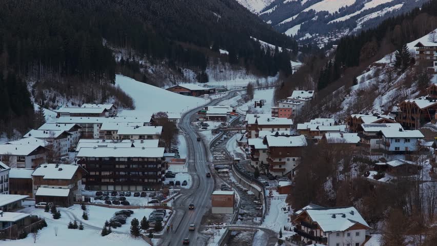 Austrian Alps landscape with snow covered village of Saalbach and cars driving on a mountain road during winter from a static elevated perspective