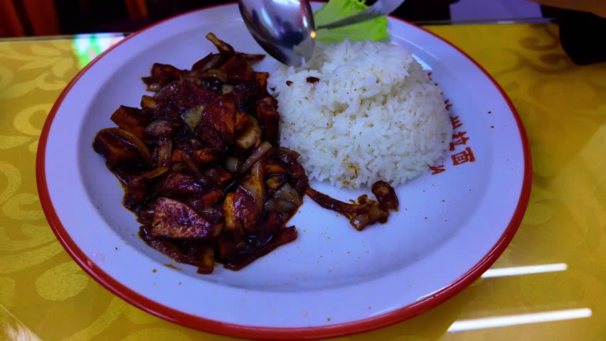 Plate of white rice with stir-fried meat and onions in dark sauce, eaten with fork and spoon on yellow tablecloth.