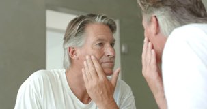 Senior man wearing white tee checking skin and jawline in bathroom mirror upon seeing reflection. Mature, selfcare, contemplative, softlighting, minimalist, home, portrait - Powered by Shutterstock - Get 15% off with code: PIKWIZARD15