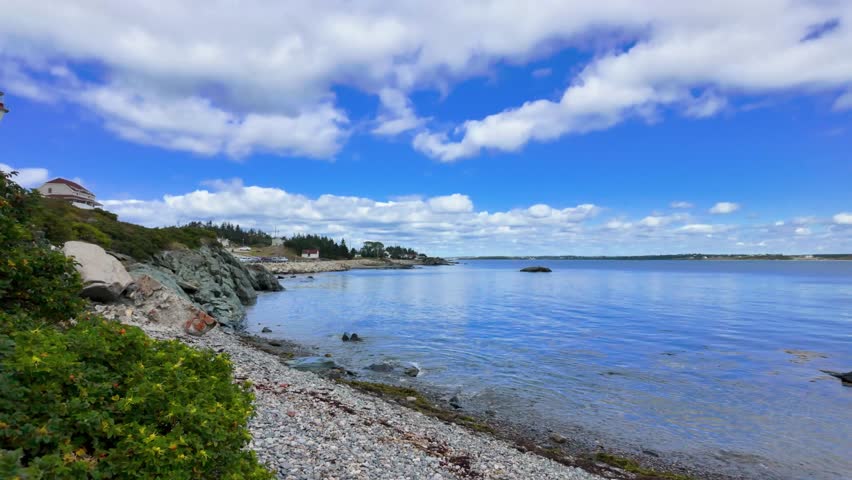 Slow motion coastal view of Cape Forchu Nova Scotia showing ocean shoreline rocks and lighthouse in bright summer daylight with clear blue sky and calm scenic atmosphere along the Atlantic coast