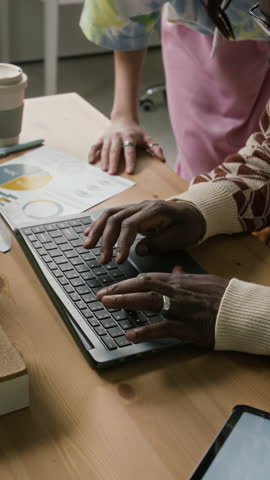 Vertical cropped shot of young woman pointing at laptop screen while senior male colleague typing on laptop during onboarding help session at desk in office