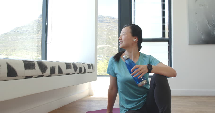 Asian woman in top starting twist on purple mat with bottle turning toward-camera smiling cooling. Yoga, stretch, seated, wellness, fitness, bright, natural