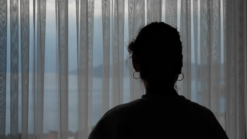 Woman opening curtain and looking at sea view. Backlit silhouette of a pensive woman opening the curtains of a hotel room window to look at the beautiful and peaceful seascape with mountains