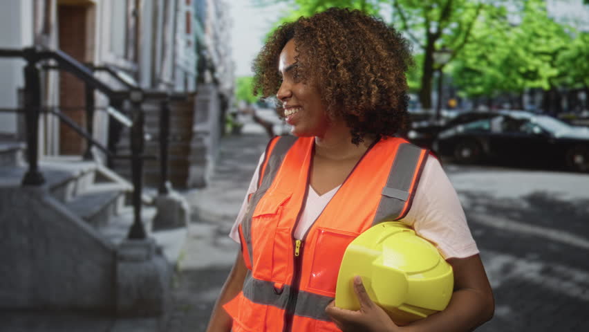 Woman engineer holding yellow helmet with hand on helmet on street wearing reflective vest and hardhat; pride teamwork confidence.