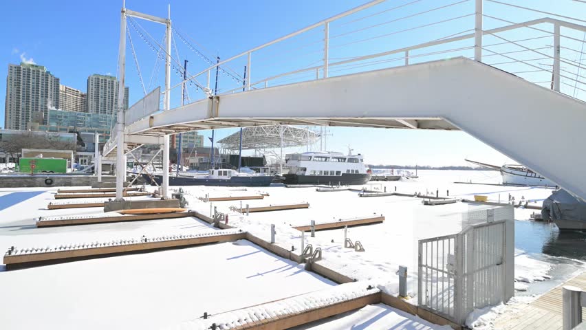 Scenic winter view of a frozen marina in downtown toronto, with snow covered docks and boats moored in the icy water under a bright, clear blue sky and modern city buildings in the background