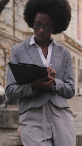Focused african woman in stylish business suit sitting with clipboard on city street. Business lady in eyeglasses writing working notes outdoors.