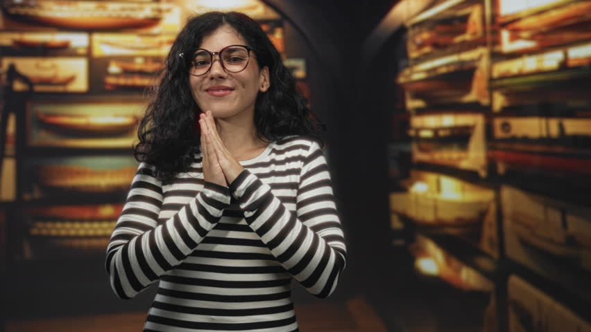 Woman with glasses claps hands softly in museum building while smiling, wearing striped top; curiosity joy.