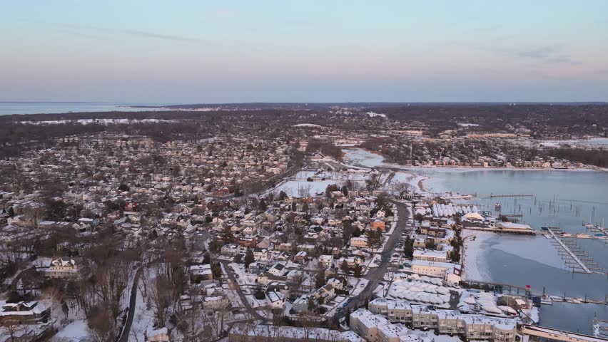 Beautiful aerial view flying over the snow covered town of port washington on long island, new york, during a calm winter sunset with a partially frozen harbor and marina