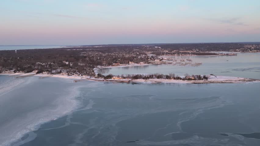 Stunning aerial perspective showing the snow covered coastline and frozen harbor of port washington during a colorful winter sunset, with ice sheets covering the calm blue water