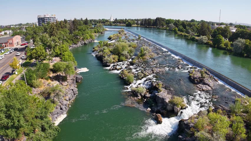 An aerial view shows a river cascading over rocky falls, with rushing water flowing through channels and trees lining the banks.
