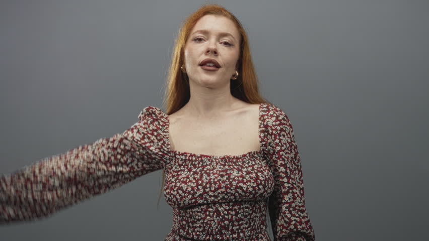 Woman covering her eyes with hand while smiling in a studio, young redhead in a floral dress showing shoulder and hoop earrings; playful joy.