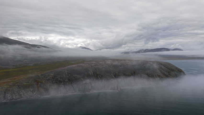 Aerial view of side tracking along Yttygran Island coast near Provideniya, Chukotka. Low fog streams over cliffs and water. Remote wilderness destination for travel projects in cloudy autumn