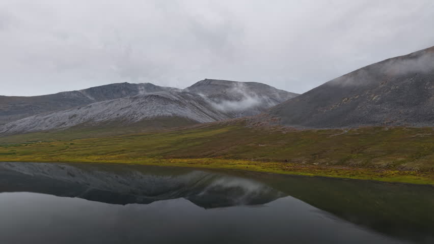aerial drone footage side tracking from lake to valley near Yttygran Island, Provideniya, Chukotka, Russia. Tundra colors and rocky slopes appear. Remote travel panorama under overcast autumn morning