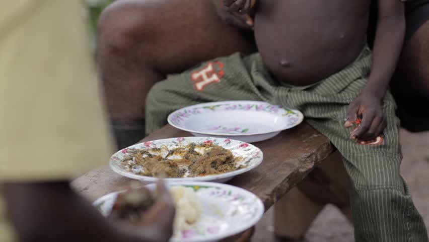 Close-up of people eating a traditional meal by hand from enamel plates on a wooden bench in rural Nigeria, with local stew and starchy “swallow” food, daily life