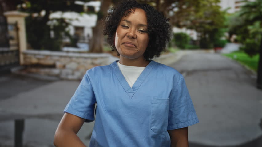 Young african american woman in blue scrubs punches air with fist on street with determined expression; victory.