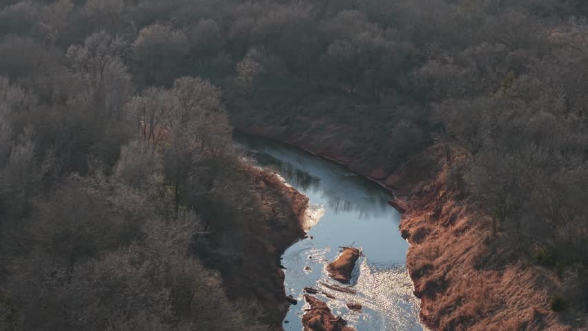 Aerial drone view of winding creek reflecting golden sunlight through dense winter forest in rural Oklahoma. River bends through dark woodland with bright seasonal light shimmering across the water surface.