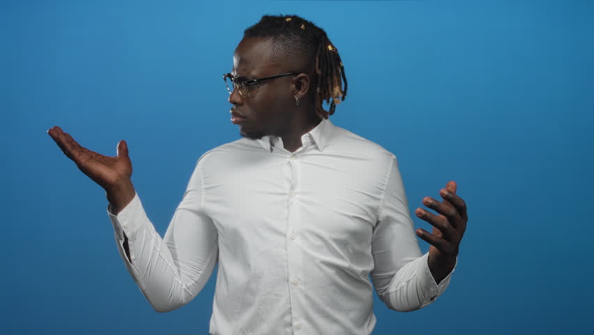 Man with palms up shrugging in blue studio wearing white shirt and glasses, dreadlocks, looking sideways; uncertainty.