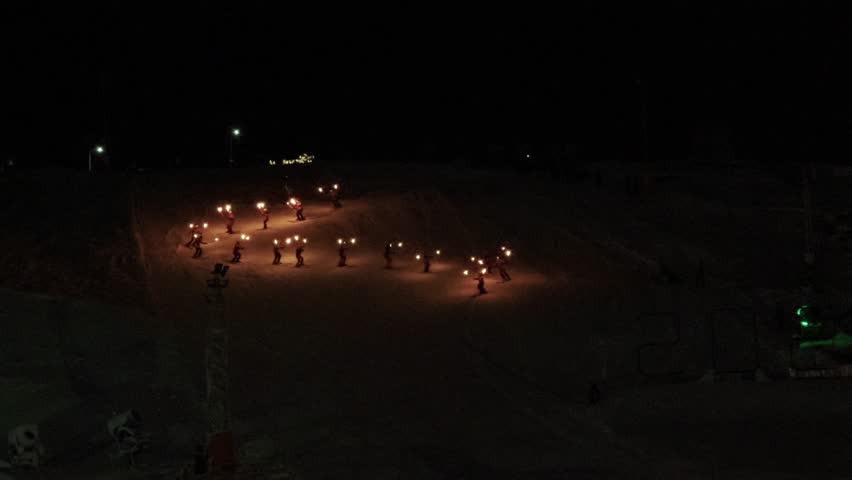 Skiers holding burning torches descending a dark ski slope during a winter evening performance, illuminating the snow