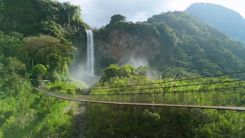 Drone shot rising to reveal Manto de la Novia waterfall plunging into lush valley with hanging pedestrian bridge over river in Baños Ecuador mountains