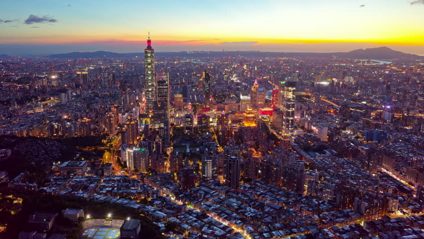 Aerial hyperlapse footage in a slow zoom-in motion at nightfall above Downtown Taipei, the vibrant capital city of Taiwan, with 101 Tower standing out amid modern skyscrapers in XinYi Commercial District, city lights dazzling under dramatic twilight sky and busy traffic trails dashing on the streets at rush hour.