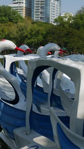 Colorful swan-shaped paddle boats and yellow duck boat docked on urban park lake with modern city skyline buildings background