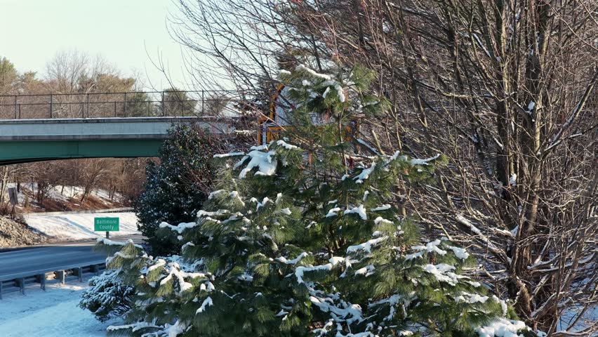 Aerial reveal shot of snowy American roadway with Maryland “Welcome” state sign, passing cars, winter trees, roadside infrastructure and calm cold-weather travel atmosphere. Wide shot.