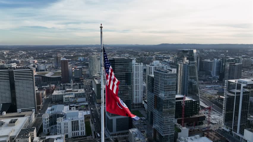 American and Tennessee flags flying above downtown Nashville, showing modern skyline, urban density and clear daylight over city center. Aerial orbit shot. Sunny day with blue sky in USA.