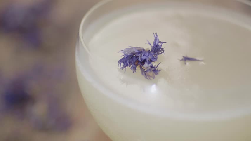 Close up of a creamy white cocktail served in a coupe glass topped with dried purple edible flowers. Refined mixology presentation showing texture and floral garnish details.