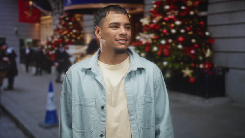 Man face looking right by decorated christmas tree on city street, wearing light denim jacket and beige t shirt; contemplation.
