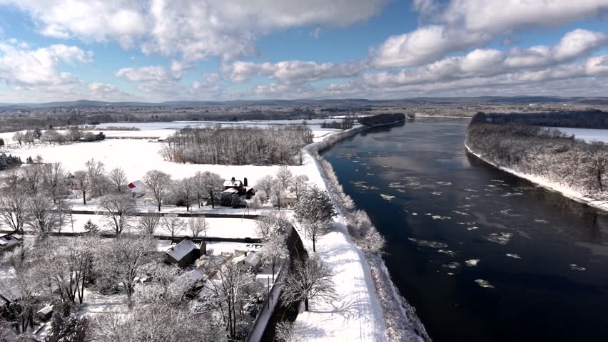 Aerial view of Connecticut River in winter, with drifting ice floes, snow-covered banks, bare trees, and rural Massachusetts farmland under bright, cold sky. Wide shot.