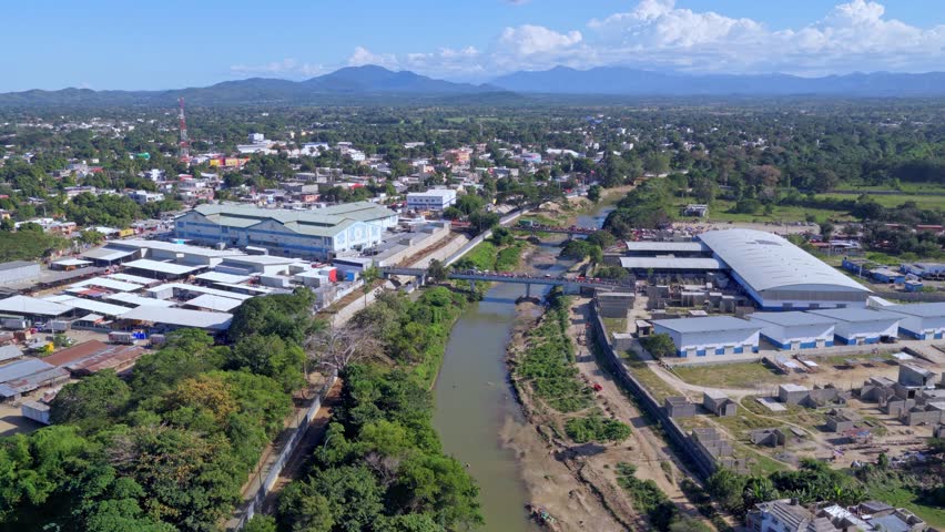 Aerial view of Dajabón border crossing between Haiti and Dominican Republic, showing bridge over river, surrounding town, industrial buildings and green landscape. Wide shot.