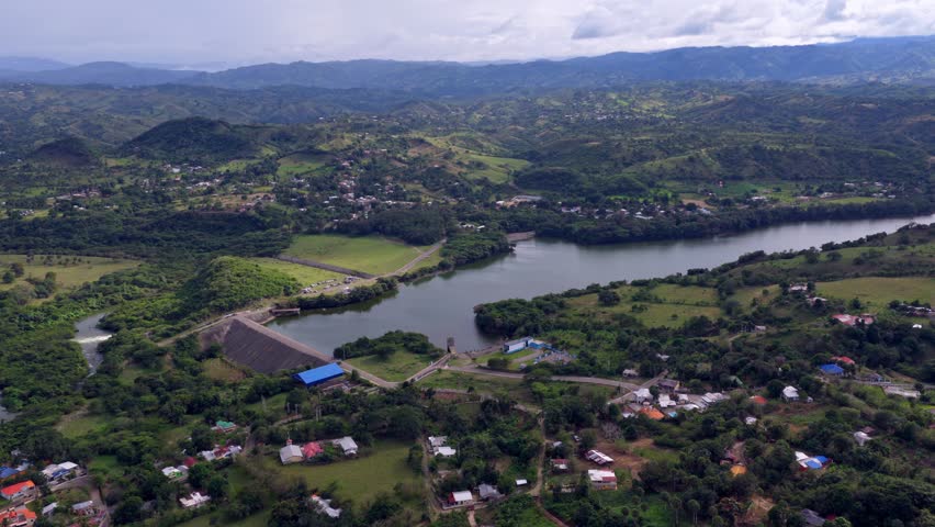 Aerial drone view of López–Angostura reservoir and dam near Santiago, Dominican Republic, showing surrounding neighborhoods, green hills, farmland and winding waterways in wide scenic landscape.