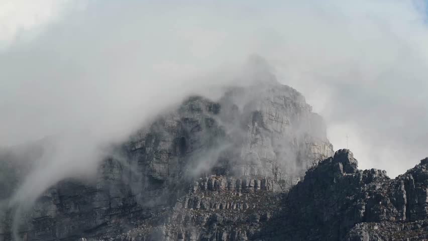 Dramatic view of Table Mountain in Cape Town, South Africa, with its iconic "Tablecloth" mist rolling over rugged rocky peaks and cliffs.