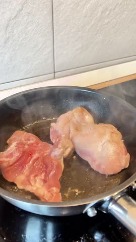 Two pieces of meat are cooking in a pan on a stove. The pan is hot and the meat is browning