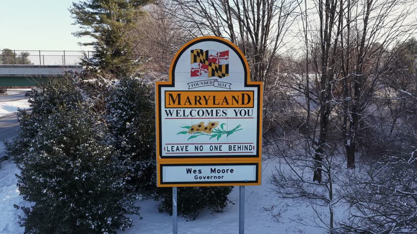 Aerial view of Maryland state welcome sign surrounded by snow-covered trees in winter, showing roadside border landmark during cold weather with clear seasonal atmosphere.