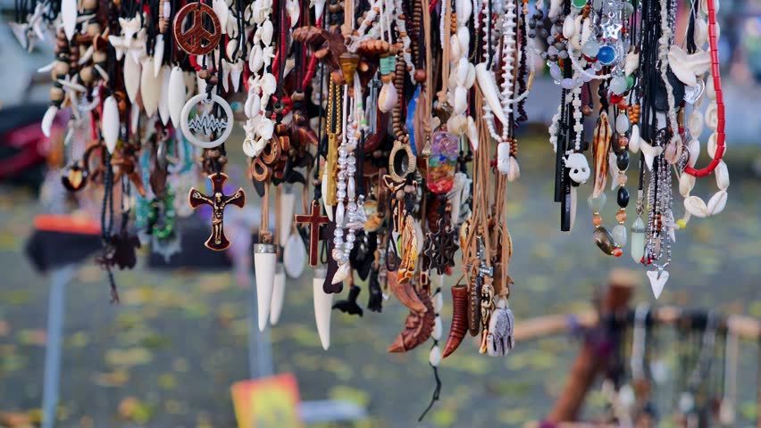 Handmade necklace collection display with wooden cross pendant, peace symbol charm, shell beads and tribal jewelry hanging at outdoor market stall, colorful boho accessory craft concept
