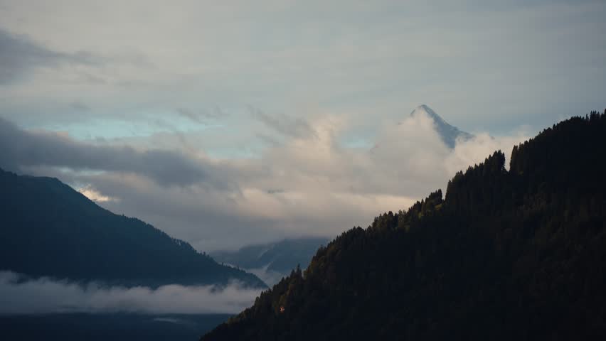 Fixed shot of Austrian alpine mountains with fast-moving clouds revealing a distant peak under an overcast sky.