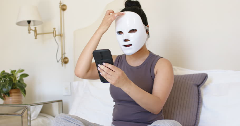 Woman is consulting smartphone on bed while adjusting white LED face mask to start skincare session. Facialdevice, selfcare, serene, bedroom, beautytech, wellness, relaxation