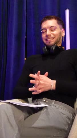Confident young man with headphones around his neck speaking and gesturing while holding a script during a podcast interview, sharing his personal story in a professional studio setting