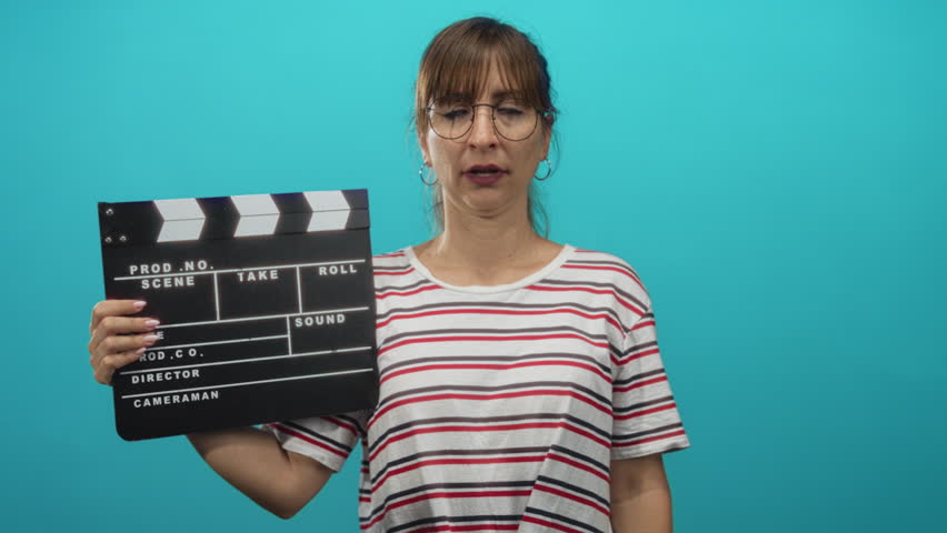 Woman holding clapperboard in teal studio wearing striped shirt and glasses; boredom filmmaking rehearsal.