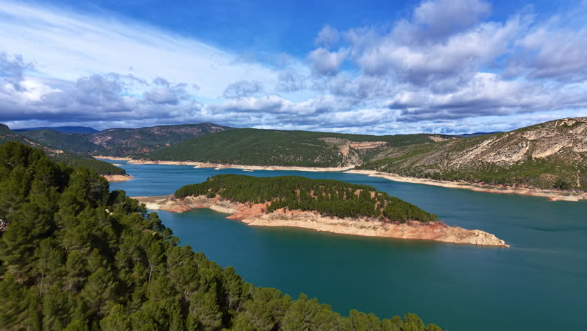 Aerial of a forested island within a mountain lake, showcasing blue water, rugged shoreline erosion lines and layered hills under dynamic cloud cover