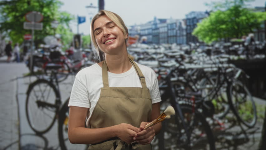 Young blonde makeup artist holding brushes, hands clasped, smiling and looking right in a studio with bicycle street backdrop; creative serenity.