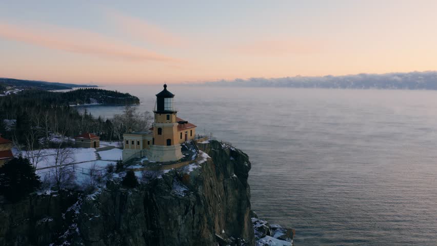 Close-up aerial orbit of the Split Rock Lighthouse lantern room and tower at sunrise with winter sea smoke in the background.