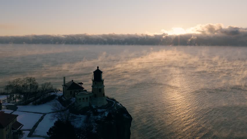 Cinematic aerial of Split Rock Lighthouse silhouetted against a golden winter sunrise with thick sea smoke on the water.
