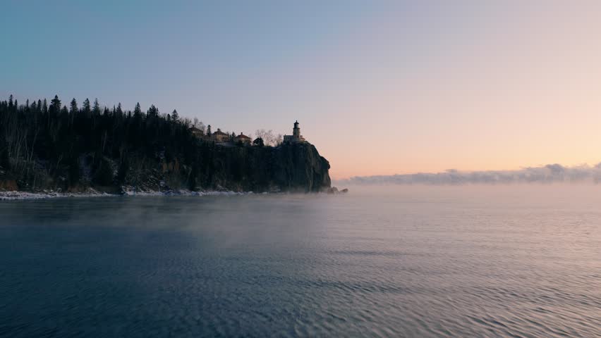 A steady low-altitude aerial shot moving across the rippling waters of Lake Superior toward Split Rock Lighthouse during a misty winter dawn.