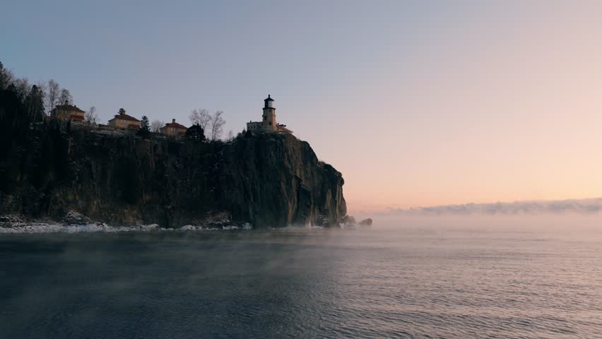 Low aerial approach over Lake Superior moving steadily toward the iconic Split Rock Lighthouse as sea smoke drifts along the cliff base.